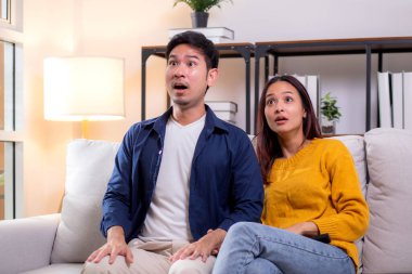 Genç Asyalı çift evdeki oturma odasında kanepede oturmuş televizyon izlerken şaşkınlıkla tepki veriyor dramatik bir sahnede duygusal tepkilerini ifade ediyor, rahat bir şekilde birlikte vakit geçiriyorlar..