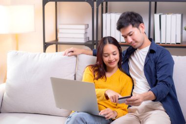 Mutlu Asyalı çift evdeki oturma odasında birlikte online alışveriş yapmak için dizüstü bilgisayar ve kredi kartı kullanıyor, kadın ve erkek gülümsüyor ve dijital yaşam tarzının keyfini çıkarıyor, uygun bir ödeme.