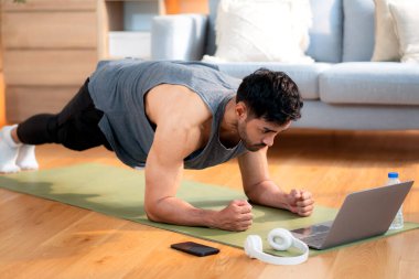 Genç Asyalı adam evde oturma odasında kalas egzersizi yaparken dizüstü bilgisayarda dayanıklılık ve motivasyonla antrenman eğitimi alırken yoga minderinde adam eğitim tahtası, dijital fitness yaşam tarzı..