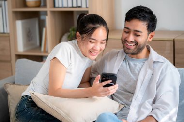 Genç Asyalı çift evdeki oturma odasında birlikte otururken rahat bir yaşam tarzı ve bağ kurma, ilişki ve boş vakit geçirme, dijital içeriği mutlu bir şekilde paylaşma ve keyif alma..
