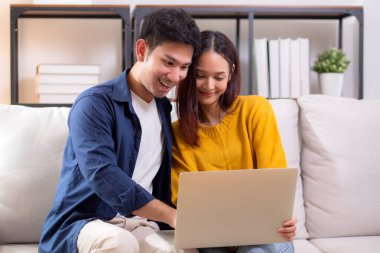 Genç Asyalı çift rahat ve rahat bir şekilde dizüstü bilgisayara bakıyor. Oturma odasındaki kanepede oturuyor. Kanepede dizüstü bilgisayar izliyor..