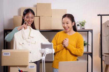 İki genç Asyalı kadın online giyim mağazasında yaşarken, evde sosyal medyada tablet ve dizüstü bilgisayarlı giysiler gösteriliyor. Girişimci çevrimiçi kıyafet satışı ve sunumu için çevrimiçi satış yapıyor..