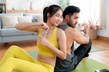 Genç Asyalı çift, evdeki antremandan sonra birlikte dinleniyorlar, yoga minderinde sırt sırta oturuyorlar egzersiz sonrası su içiyorlar, ferahlatıcı ve iyi hissediyorlar, mola veriyorlar..