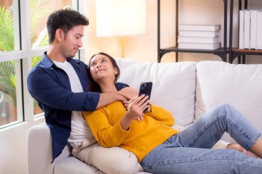 Mutlu genç Asyalı çift, evlerinde rahat bir koltukta rahat rahat otururken akıllı telefon kullanıyorlar. Akıllı telefonları dinlenmek, şefkat ve modern ev hayatı için kullanıyorlar..