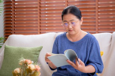 Asyalı son sınıf öğrencisi bir kadın kanepede oturmuş kitap okuyor. Oturma odasında dinleniyor. Yaşlı bir kadın hafta sonu rahat, hobi ve eğlence dolu bir kanepede edebiyat okuyor..