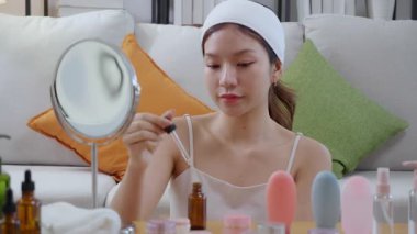 Güzel, genç Asyalı bir kadın ellerine losyon sürüyor. Evdeki aynaya bakıyor. Ellerine gençleştirme için güzellik serumu, kendine bakım, cilt bakımı ve sağlık için makyaj yapıyor..