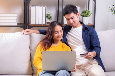Mutlu Asyalı çift evdeki oturma odasında birlikte online alışveriş yapmak için dizüstü bilgisayar ve kredi kartı kullanıyor, kadın ve erkek gülümsüyor ve dijital yaşam tarzının keyfini çıkarıyor, uygun bir ödeme.