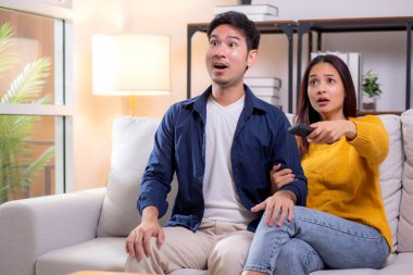 Genç Asyalı çift evdeki oturma odasında kanepede oturmuş televizyon izlerken şaşkınlıkla tepki veriyor dramatik bir sahnede duygusal tepkilerini ifade ediyor, rahat bir şekilde birlikte vakit geçiriyorlar..