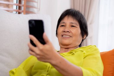 Üst düzey Asyalı kadın kanepeye oturmuş videolu telefon bekliyor. Oturma odasında. Yaşlılar sosyal medyayı internet üzerinden sohbet etmek için kullanıyor..