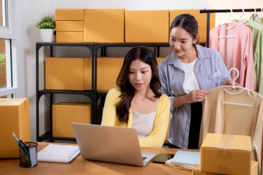 İki genç Asyalı iş kadını dizüstü bilgisayar, sevkiyat ve kargo için müşteri kıyafetlerini kontrol ediyor, çevrimiçi satıcı ve mağaza ile perakende satış, başlangıç veya küçük işletme, nakliye ve envanter.
