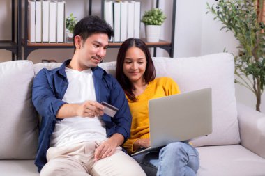 Mutlu Asyalı çift evdeki oturma odasında birlikte online alışveriş yapmak için dizüstü bilgisayar ve kredi kartı kullanıyor, kadın ve erkek gülümsüyor ve dijital yaşam tarzının keyfini çıkarıyor, uygun bir ödeme.