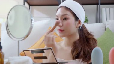 Güzel, mutlu, Asyalı bir kadın oturma odasında aynaya bakarak makyaj yapıyor. Güzel bir kadın makyaj yapıyor. Cazibe ve zarafet için yüz makyajı yapıyor..