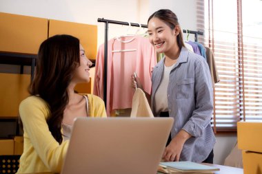 İki genç Asyalı iş kadını dizüstü bilgisayar, sevkiyat ve kargo için müşteri kıyafetlerini kontrol ediyor, çevrimiçi satıcı ve mağaza ile perakende satış, başlangıç veya küçük işletme, nakliye ve envanter.