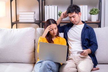 Dizüstü bilgisayara bakan Asyalı çift, oturma odasında mali sorun yaşarken, dizüstü bilgisayar kullanan kadın ve erkek kredi kartını üzgün bir şekilde tutarken, ifade bütçesi endişe ve masraflı bir çift olarak vurguladı..