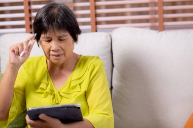 Kıdemli Asyalı kadın kanepeye oturmuş dijital tablet kullanıyor. Oturma odasında dinleniyor, yaşlılar tablete oturmuş düşünceli, rahat bir hayat tarzı ve iletişim kuruyor..