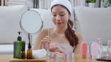 Güzel Asyalı bir kadın, oturma odasında aynaya bakarken kırışıklıklar için yüzüne krem sürüyor. Güzel bir kadın, yanağına nemlendirici sürüyor. Hijyen, cilt bakımı ve güzellik için..