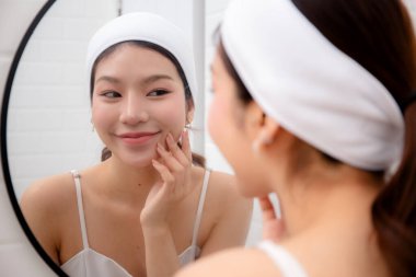 Güzel, genç Asyalı kadın teni banyodaki aynaya bakarken yüzünde kırışıklıkları gülümsemeyle, pürüzsüz teniyle ve tatmin olmuş cildiyle kontrol eden kadın, güzellik ve cilt konsepti..