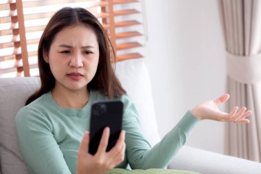 Genç Asyalı kadın evdeki akıllı telefona bakarken kanepede oturmuş endişeli ve stresli bir şekilde, mutlu olmayan ve endişeli bir kadın akıllı telefondan, yaşam tarzından ve iletişimden izliyor..