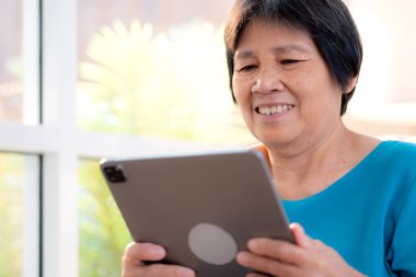 Kıdemli Asyalı kadın evdeki oturma odasında tablet okurken, internet bağlantısı ve dijital yaşam tarzı, sosyal medya için tablet izlerken, uzaktan öğrenirken, sohbet robotu veya yapay zeka kullanırken, kanepede dinleniyor..