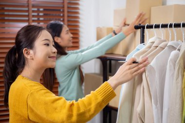 İki genç Asyalı kadın, envanter, çevrimiçi satıcı, küçük işletme ve taşımacılık ile belge üzerinde kontrol ve planlama, satıcı veya girişimci ofis teslim için stok giysileri kontrol ve planlama.