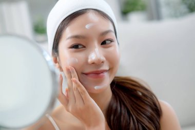 Güzel Asyalı bir kadın, oturma odasında aynaya bakarken kırışıklıklar için yüzüne krem sürüyor. Güzel bir kadın, yanağına nemlendirici sürüyor. Hijyen, cilt bakımı ve güzellik için..