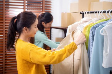 Hazırlık teslimatı için mağazada kıyafet düzenleyen iki genç Asyalı kadın, satıcı seçici giysiler, perakende satış için seçici giysiler, teslimat için mal, küçük işletme.