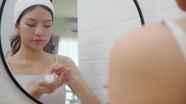 Genç ve güzel Asyalı bir kadın banyoda aynaya bakarken cildine krem sürüyor ya da losyon sürüyor. Tedavi için nemlendirici sürüyor. Yüz hatları, güzellik ve cilt bakımı için..