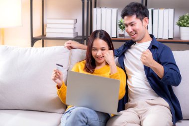 Mutlu Asyalı çift evdeki oturma odasında birlikte online alışveriş yapmak için dizüstü bilgisayar ve kredi kartı kullanıyor, kadın ve erkek gülümsüyor ve dijital yaşam tarzının keyfini çıkarıyor, uygun bir ödeme.