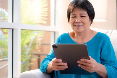Kıdemli Asyalı kadın evdeki oturma odasında tablet okurken, internet bağlantısı ve dijital yaşam tarzı, sosyal medya için tablet izlerken, uzaktan öğrenirken, sohbet robotu veya yapay zeka kullanırken, kanepede dinleniyor..
