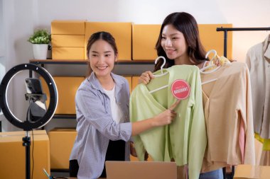 İki genç Asyalı kadın online kıyafet mağazasında canlı yayında canlı yayında sosyal güvenlik telefonuyla kıyafetlerini gösterirken, girişimci online kıyafet satışı ve sunumu için online satıcı,.