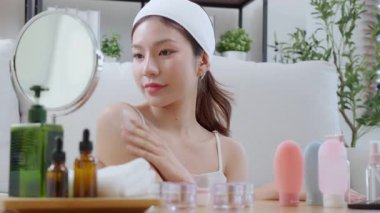 Güzel, genç Asyalı bir kadın, oturma odasında aynaya bakarken krem ya da losyon sürüyor. Omzunda, cildinde ve sağlığında gençleşme için cilt bakımı yapan mutlu, güzel bir kadın..