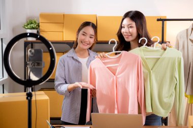 İki genç Asyalı kadın online kıyafet mağazasında canlı yayında canlı yayında sosyal güvenlik telefonuyla kıyafetlerini gösterirken, girişimci online kıyafet satışı ve sunumu için online satıcı,.