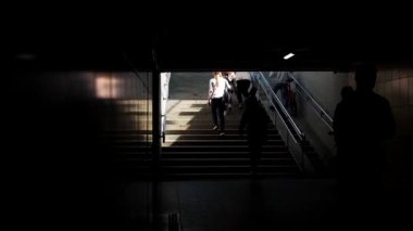 Altgeçitten arka planda bir grup insan çıkıyor. Yayalar metro merdivenlerinden güneş ışığına doğru ilerliyorlar. Şehir hayatı ve koşuşturma. İnsanların gölgeleri