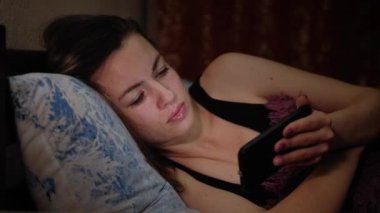 Genç beyaz kız gece yatakta yatarken akıllı telefon kullanarak video izliyor. Güzel bir kadın akşamları yatak odasında yatarken alet ekranına bakar..