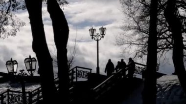 Arka planda, bulutlu güzel bir gökyüzünün arka planına karşı merdivenlerden inen insanların ana hatları. Arka planda bir kalabalık parkta yürüyor. Kış ya da ilkbahar.