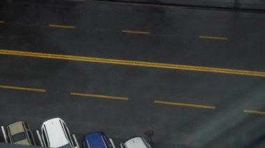 Şehir yolu ve park yerindeki ofis penceresinin üst görüntüsü. Arabalar cadde boyunca geçer, insanlar yolun karşısına geçer. Alçak açı atışını kapat. Şehir caddesinde sarı yol işaretleri. Şehir tarzı