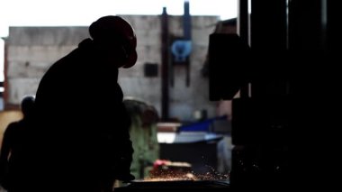 Arka aydınlatma işçisi bir levhayı öğütücüyle temizliyor. Arka planda metali profesyonel bir aletle kesen bir adamın dış hatları. Ağır sanayi metalürji fabrikası.