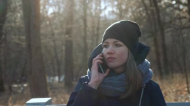 Parktaki bankta oturan genç beyaz bir kız palto giyiyor, telefonda konuşuyor ve gülümsüyor. Güzel beyaz kadın bahar başında ya da sonbaharın sonunda şehir parkında oturur. Tam HD 50 peni video kaydı