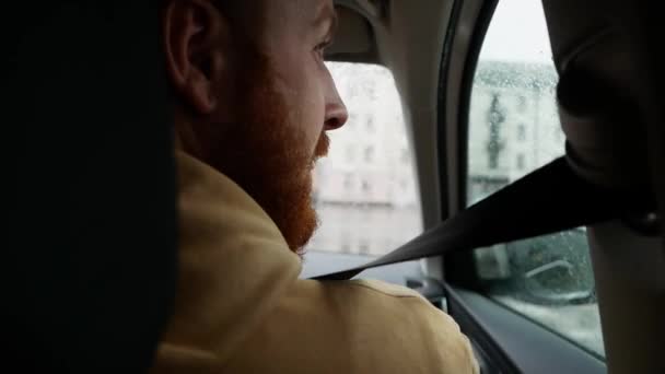 一个留着红胡子的白人坐在汽车的前座上 系着安全带 一个男人在雨天开车 从后座看到一个乘客 — 图库视频影像