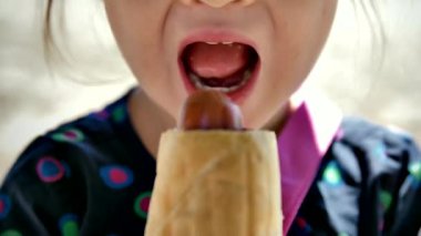 Sosisli yiyen tatlı küçük bir kızın ağzının yakın çekimi. Komik çocuk fast food yer ve dudaklarını yalar. Çocuk aç gözlülükle sokakta yürürken bir topuzu ısırıyor..