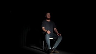 Sakallı bir adam karanlık bir odada, açık bir kapı ya da pencerenin ışığıyla aydınlatılmış bir sandalyede düşünceli bir şekilde oturur. Adam dramatik sahneler, yalnızlık, umut.