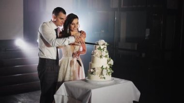 Yeni evliler şenlikli bir ziyafet sırasında büyük, güzel bir düğün pastası kestiler. Gelin ve damat bir düğün tatlısını paylaşıyor. Beyaz gül çiçekleriyle süslenmiş beyaz pasta..