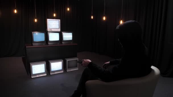 许多老式坏电视机前的扶手椅上坐着一个黑帽陌生人 并按下电视遥控器上的一个按钮 假新闻 媒体操纵和僵尸电视的概念 Glitch — 图库视频影像