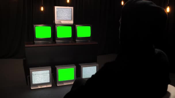 黒フードの見知らぬ人は キーとカットのためのグリッチ静的ノイズと緑のクロマキー画面を示すテレビのスタックの前に座っています メディア宣伝と操作の概念 情報戦争 — ストック動画