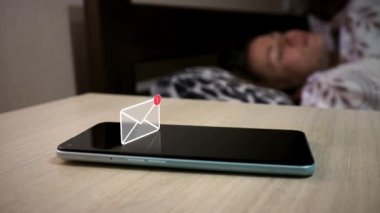 Komodinin üzerinde duran cep telefonunda kısa mesaj bildirimi görülüyor. Telefonun üstündeki hologram gibi animasyon simgesi. Yatakta uyuyan kadının arka planında cep telefonu var.