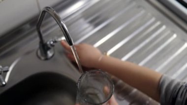 Yavru kız, akan su filtresi olan bir musluktan bir bardak temiz filtrelenmiş su dolduruyor. Çocuk içmek için su topluyor. Su dengesi ve susuzluk.