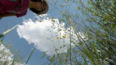 Küçük sarı çiçeklerin üzerine eğilen, onları koklayan ve sonra onları koparan genç kız portresi. Çocuk, gür bulutlu mavi gökyüzüne karşı güzel çiçekler topluyor. Aşağıdan geniş açı.