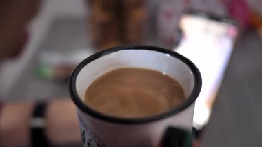 Bir kadın sıcak bir içeceği soğutmak için üfler. Soğuk mevsimde bir kız sıcak bir içecek içer, kahvaltıda akıllı telefonuyla video izler ve düşünür..