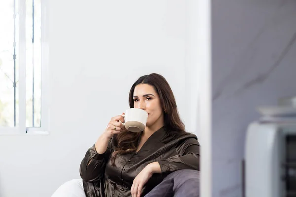 美しい若いヒスパニック系の女性がコーヒーを飲みながら家に座って ストックフォト