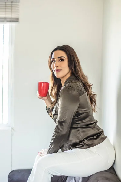 stock image sensual beautiful hispanic feminine woman sitting on sofa with cup of coffee. relaxed latin woman watching the cam